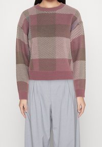 Pull multicolore avec un motif à carreaux dans des nuances de rose, marron et gris. Poignets et ourlet côtelés ; coupe oversize. Associé à un pantalon gris.