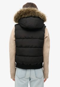 Schwarze Steppweste mit abnehmbarer Kapuze und Kunstfellbesatz. Die Weste hat einen gerippten Saum und wird mit einem beigen Pullover kombiniert.