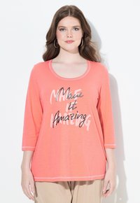 Korallenfarbener Langarmtop mit Rundhalsausschnitt und kontrastierenden Nähten. Auf der Vorderseite steht "Make it Amazing" in schwarzem und grauem Text.