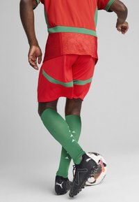 Röd atletisk tröja och shorts med gröna detaljer; gröna knästrumpor med vita triangelmönster; svarta fotbollsskor; en fotboll.