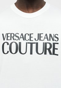Κοντινό πλάνο ενός ατόμου που φοράει λευκό μπλουζάκι με έντονα μαύρα γράμματα «VERSACE JEANS COUTURE» στο στήθος.