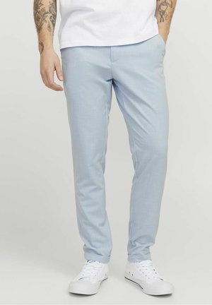 Homme portant un pantalon slim bleu clair, des baskets blanches et une chemise blanche avec les bras tatoués visibles, debout les mains dans les poches.