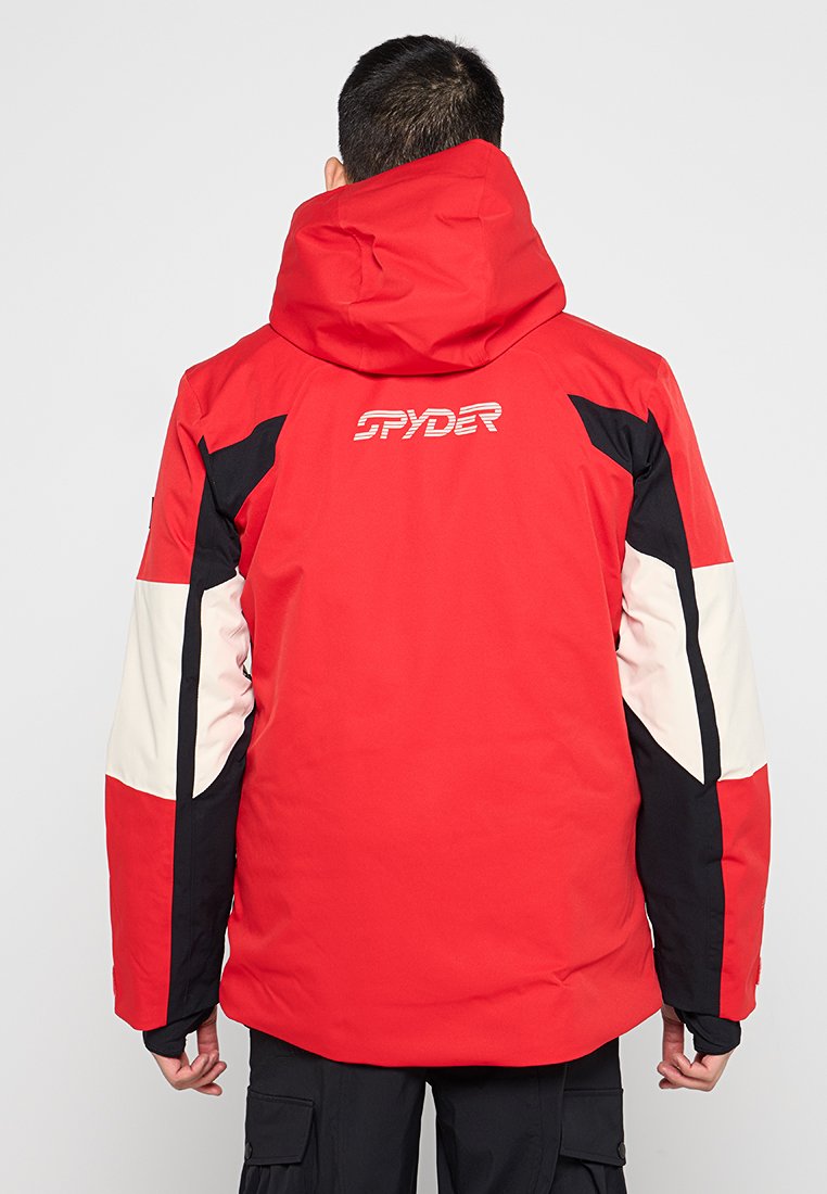 Mann trägt eine rote, schwarze und weiße Spyder-Skijacke mit Kapuze, von hinten vor einem schlichten grauen Hintergrund betrachtet.