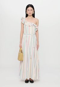 Mulher vestindo um vestido maxi listrado multicolorido de um ombro só com detalhe de folhos, segurando uma bolsa de vime e usando sapatilhas pretas.