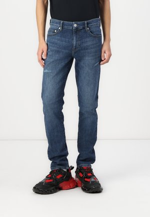 Jeans din denim de culoare albastru mediu, cu un design ajustat, prezentând ușoare uzuri, cinci buzunare și o închidere clasică cu buton. Purtați cu teniși negri-roșii.