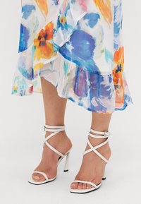 Robe florale avec des motifs bleus, oranges et jaunes ; ourlet à volants superposés ; associée à des sandales à talons hauts blanches avec un bout carré.