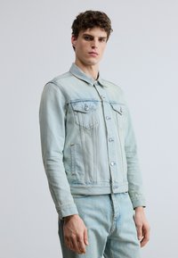 Ljus denimjacka med krage, två bröstfickor och knäppningar med knappar. Blekt blå färg med en lätt sliten struktur.