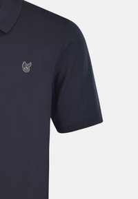 Marineblå pikétröja med korta ärmar, tillverkad i bomull. Har en subtil broderad logotyp på vänster bröstområde och en klassisk krage.