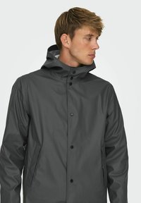 Jachetă impermeabilă gri cu guler înalt, închidere cu nasturi și buzunare laterale. Textură netedă cu un design minimalist.