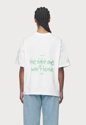 Persoon met gevlochten haar die een wit T-shirt draagt met groene tekst "the night ones won't leave" en lichtblauwe spijkerbroek, gezien van achteren.