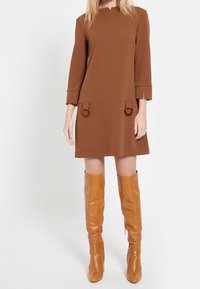 Brun knälång klänning med trekvartslånga ärmar och cirkulära detaljer, i kombination med högblanka tan stövlar upp till låret. Slät textur.