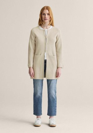 Kvinde med rødt hår står iført en beige lang cardigan over en hvid top, blå jeans og hvide sneakers mod en ensfarvet baggrund.