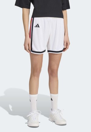 Person trägt weiße Adidas-Sportshorts mit schwarzen Abschlüssen und Seitenstreifen, weiße Adidas-Socken und weiße Sneakers, steht mit entspannten Armen.