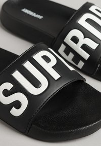 Zwarte slipper sandalen met brede rubberen bandjes met gedurfde witte "SUPER" letters. Gevoerde zwarte voetbed voor comfort. Plat zool ontwerp.