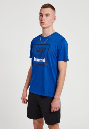Młody mężczyzna w niebieskiej koszulce z logo hummel, czarnych szortach i srebrnym łańcuszku na szyi, stojący na jednolitym jasnym tle.