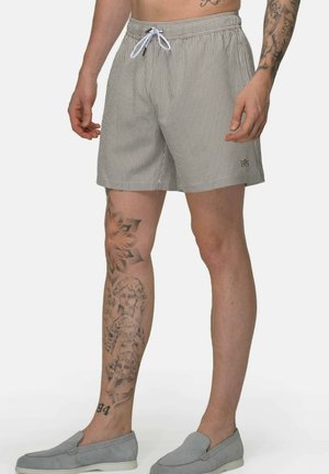 Man draagt gestreepte shorts met trekkoord en grijze instapschoenen, met gedetailleerde tatoeages zichtbaar op het linkerbeen en de rechter onderarm.