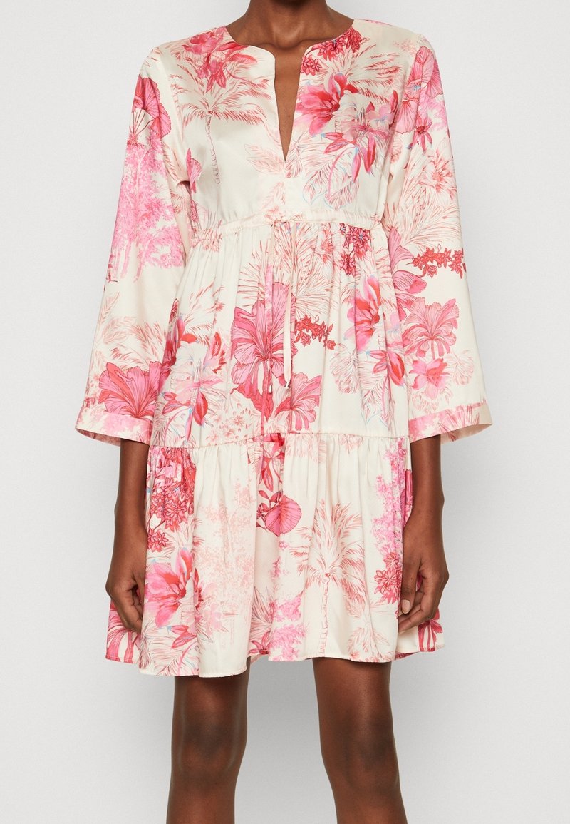 Femme portant une robe crème mi-longue avec un imprimé floral tropical rose et rouge, manches longues et un nœud devant à la taille.