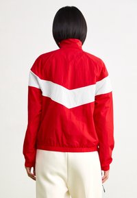 Persona de espaldas vestida con una chaqueta roja que tiene una franja blanca en forma de chevron en la parte de atrás y pantalones blancos, contra un fondo liso.