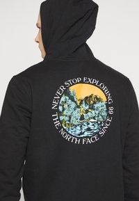 Personne portant un sweat à capuche noir montrant un graphique circulaire avec des montagnes, des arbres, une rivière, un coucher de soleil et le texte « Never Stop Exploring The North Face Since 66 ».