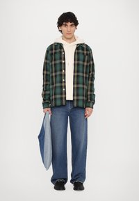 Grønn rutete flanellskjorte med gule detaljer, lagt over en beige hettegenser, kombinert med blå vide bukser og svarte sko, holding en denim tote.