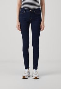Jeans ajustados de color azul oscuro hechos de mezclilla, con bolsillos frontales, cremallera y costuras visibles. Combinados con zapatillas gris y blancas.