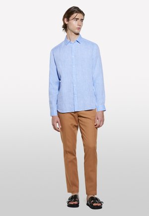 Camicia azzurra chiara a maniche lunghe con bottoni frontali, abbinata a pantaloni beige. Il modello indossa sandali neri con dettaglio a fibbia. Tessuto dalla consistenza liscia.