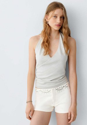 Jeune femme aux longs cheveux ondulés portant un haut sans manches gris clair style dos nu et un short blanc avec des détails cloutés, debout contre un mur uni.