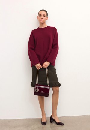 Burgundowy oversize sweter, oliwkowa spódnica z frędzlami, burgundowa torebka na ramię z logo oraz pasujące szpilki, stojące na gładkiej podłodze.