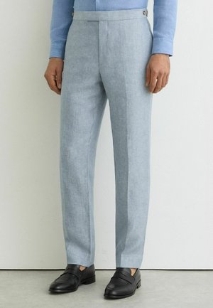 Uomo che indossa pantaloni su misura azzurri, mocassini neri in pelle e una camicia blu a maniche lunghe, in piedi contro un muro bianco.