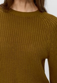 Maglione lavorato a maglia color senape con una finitura testurizzata, caratterizzato da un collo rotondo e polsini a coste, che mostra una vista ravvicinata del tessuto.