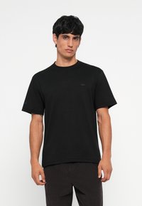 T-shirt en coton noir avec col rond, manches courtes et un petit logo noir sur la poitrine. Comprend un texte bleu sur une manche.
