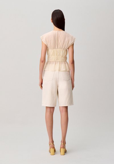 Donna con lunghi capelli scuri che indossa un top beige trasparente arricciato, pantaloncini color crema alla lunghezza del ginocchio e sandali gialli con tacco, in piedi davanti a uno sfondo semplice.