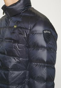 Giacca puffer navy con colletto alto, dotata di chiusura con zip, baffle orizzontali e una patch logo nera sulla spalla.
