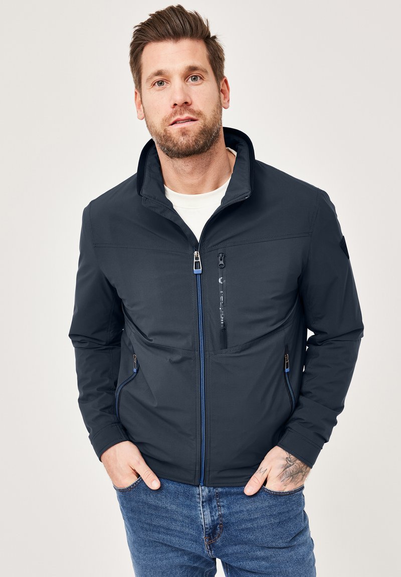 Navy blue softshell jacket with stand collar, zippered pockets, and contrasting blue accents. Fabric appears smooth and water-resistant.