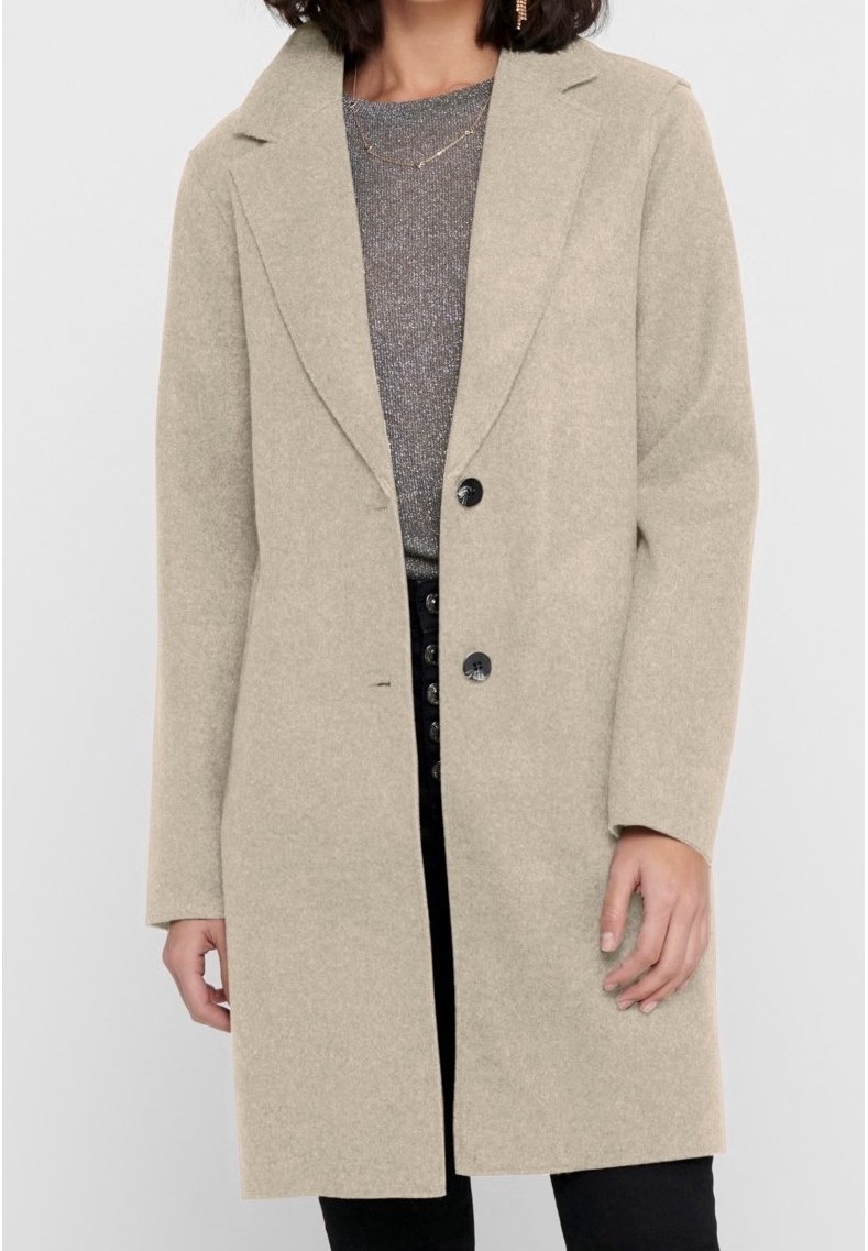Femme portant un long manteau en laine beige par-dessus un haut gris et un pantalon noir avec de grands boutons, debout devant un fond uni.