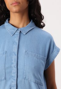 Camisa de denim azul claro com gola, mangas curtas dobradas e dois bolsos no peito, com uma textura suave.
