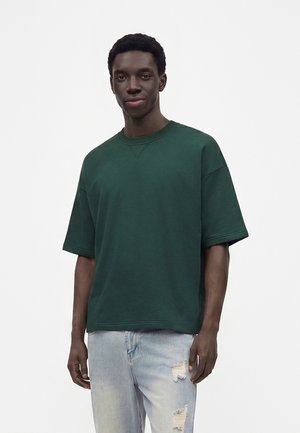 Jeune homme aux cheveux courts et bouclés portant un sweat-shirt ample à manches courtes vert foncé et un jean bleu clair déchiré, debout sur un fond uni.