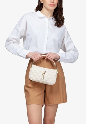 Donna con camicia bianca abbottonata e pantaloncini color sabbia, che tiene davanti a sé una piccola borsa a tracolla bianca imbottita con logo dorato e manico.