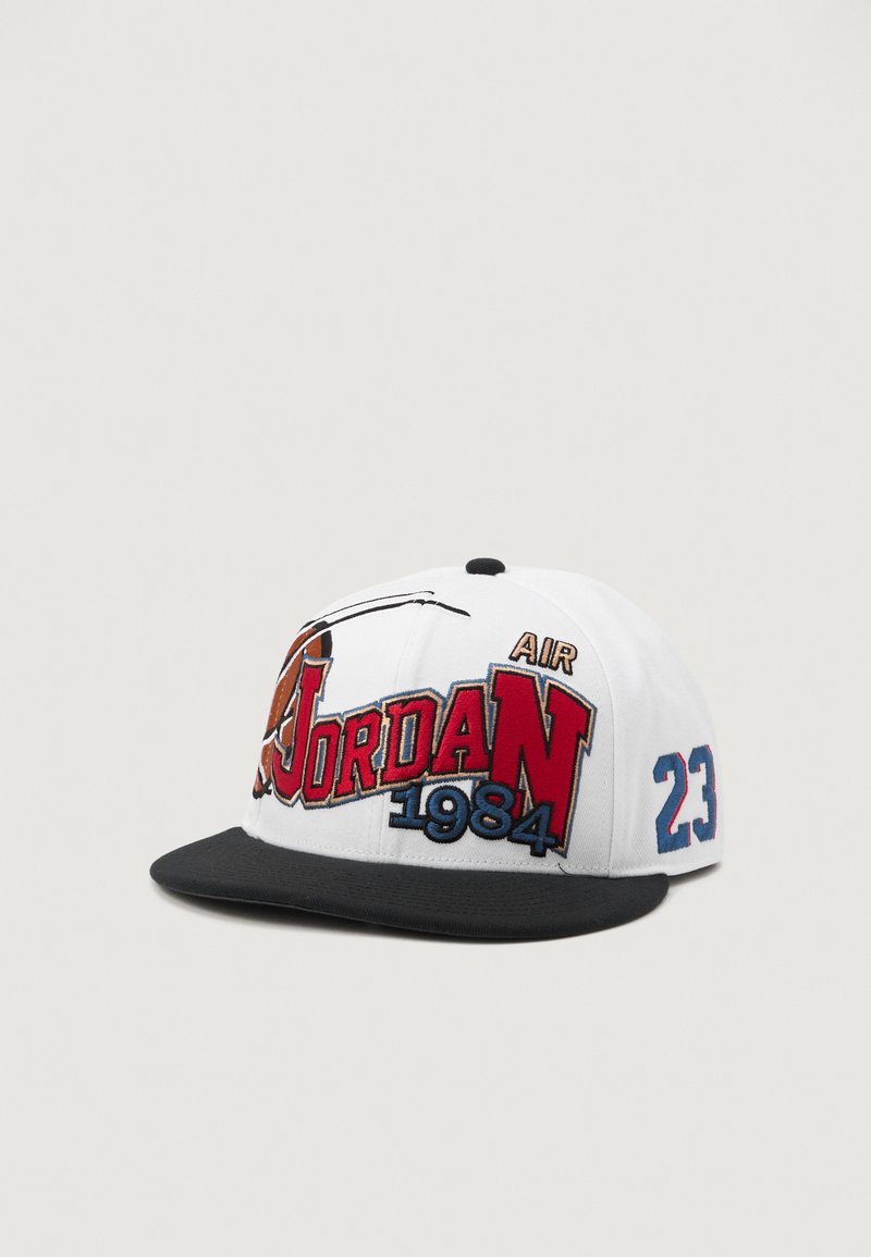 Fehér baseball sapka fekete karimával. Piros és kék hímzett "Jordan" felirattal, egy kosárlabda grafikával, valamint "1984" és "23" felirattal az oldalon.