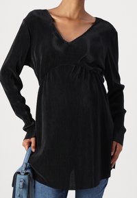 Mustassa ribattua pitkähihaista toppia, jossa on V-mallinen kaula-aukko, kerätty vyötärö ja pehmeä tekstuuri, yhdistettynä sinisiin farkkuihin ja vaaleansinsiseen laukkuun.