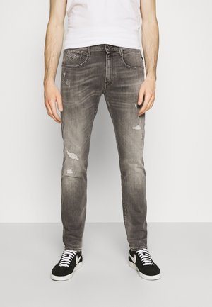 Man wearing faded black slim-fit jeans with distressed patches, white T-shirt, and black sneakers with white laces and soles.