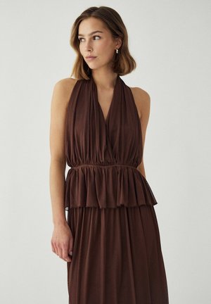 Õlgadeni juustega naine, kes kannab varrukateta pruuni kaelasiduvat kleiti, millel on voldiline vöökoht, vaatab vasakule ühevärvilise tausta ees.