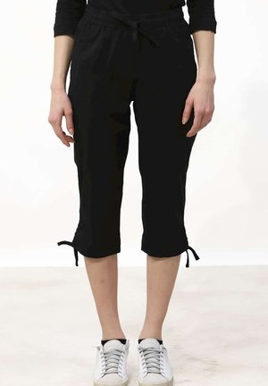 Person trägt schwarze, gekürzte Hosen mit Bindebändern am Saum und weiße Sneakers, steht auf einem hellen Boden vor einem schlichten Hintergrund.