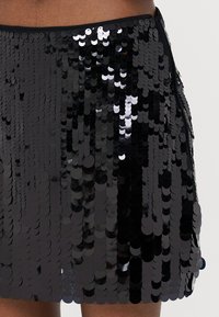 Juodas blizgučiais padengtas mini sijonas su tiesiu krovu, pasižymintis apvaliais, blizgiais diskas, glaudžiai išdėstytais tekstūrai ir vizualiniam interesui.