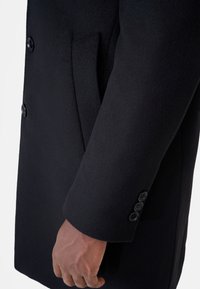 Svart ullmantel med en slät textur, sidoficka och tre-knapps manschettdetalj. Renskuren design med skräddarsydd passform.