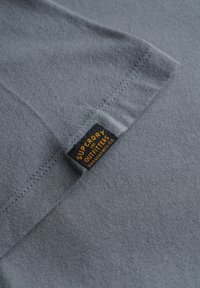 Blauw-grijze stofdetail met gestikte zoom en een klein zwart label met de tekst "Superdry GBR Outfitters Garment Mfg Co." in oranje letters.
