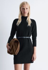 Schwarzes, strukturiertes Kleid mit taillierter Silhouette, hohem Kragen und kurzen Ärmeln, in der Taille mit einem blauen Gürtel geschnallt. Braune Tasche in der Hand.