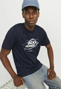 Navyblauwe katoenen t-shirt met een witte grafische print met de tekst "JACK & JONES." Het model draagt een lichtblauwe pet en een lichte spijkerbroek.