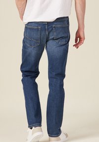 Dunkelblaue Jeans aus Denim mit geradem Bein, ausgestattet mit Gesäßtaschen und einem sauberen Abschluss am Saum. Getragen mit weißen Schuhen.
