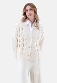 Strickjacke in Creme mit einem strukturierten Rautenmuster, V-Ausschnitt und Knopfverschluss. Kombiniert mit einem weißen Hemd und passenden Hosen.